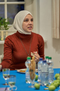 2020-10-14-SBHeV-7Jubiläum__DSC3410_2048px Eine junge Frau mit Kopftuch sitzt an einem Tisch, auf dem einige Wasserflaschen zu sehen sind. Sie trägt ein rotes Oberteil und schaut aus dem Foto heruas.