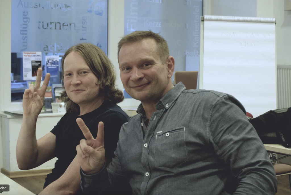 Foto – Siena 14 Eine Frau und ein Mann während einer Gesprächsrunde von Sprachbrücke-Hamburg e.V. lächeln in die Kamera und zeigen das Victory-Symbol mit ihrer jeweils linken Hand.