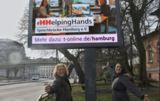 Annja Haehling von Lanzenauer und Susanna Jordanovic von Sprachbrücke-Hamburg e.V. vor einer Stroer-Werbetafel und der #HHelpingHands-Werbung