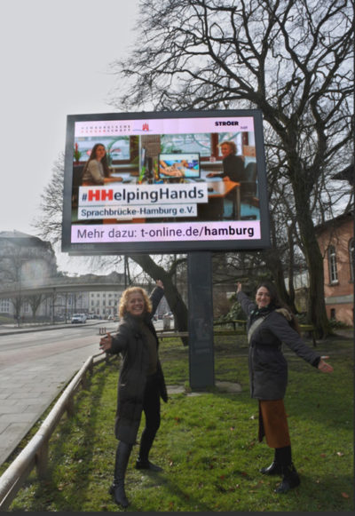 2023-03-08-Helping-Hands Annja Haehling von Lanzenauer und Susanna Jordanovic von Sprachbrücke-Hamburg e.V. vor einer Stroer-Werbetafel und der #HHelpingHands-Werbung