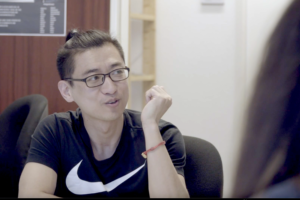 AWO Gesprächsrunde Ein junger Mann asiatischer Herkunft mit Brille, Kurzarm-Shirt und zurückgebundenen Haaren sitzt an einem Tisch und schaut zur Seite im Gespräch mit einer oder mehreren Personen