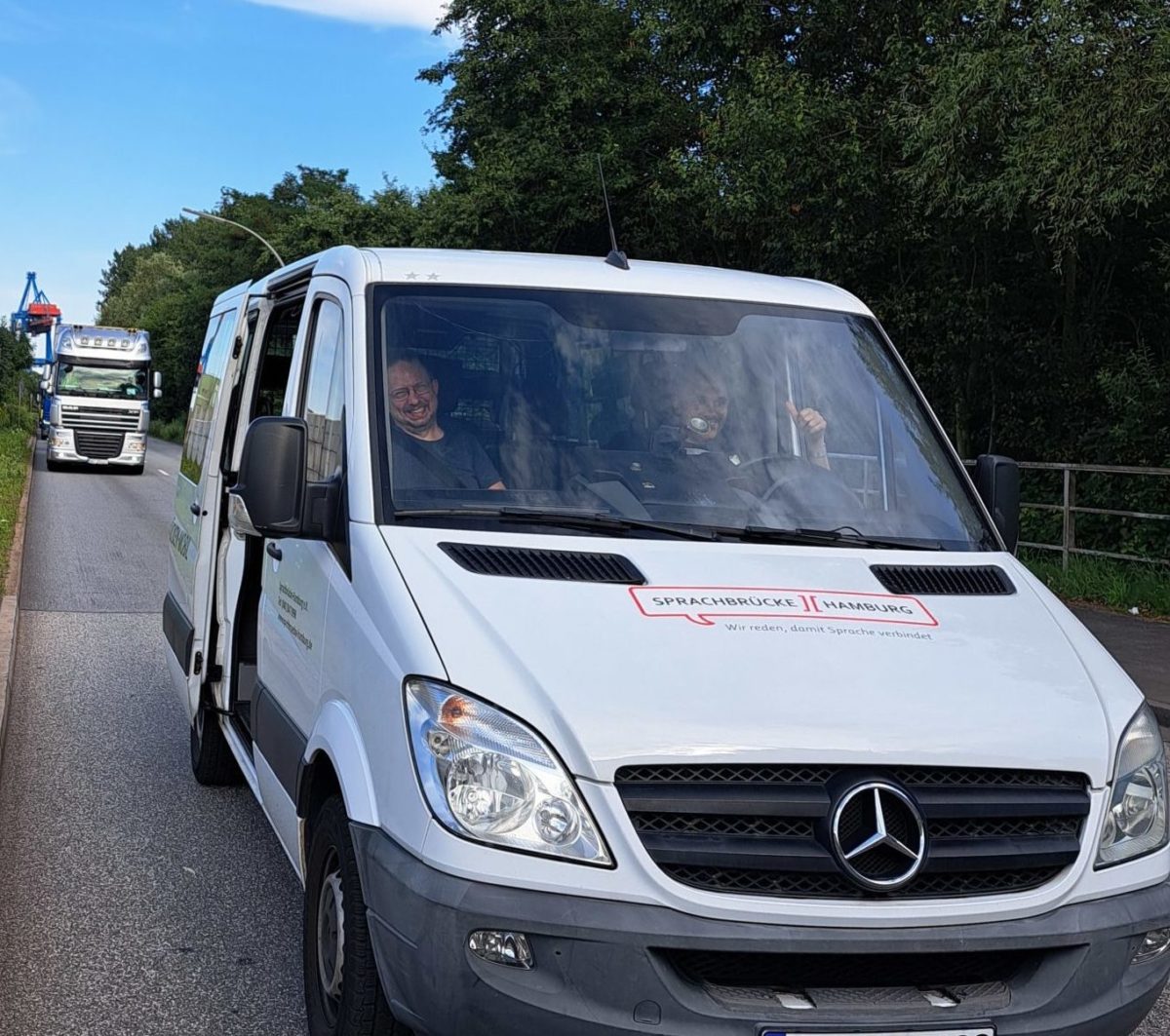 SB-Mobil in Neuenfelde_2024-07-15 Ein weißer Transpoerter steht mit 2 Fahrern in einer langen Einfahrt. Daneben ist eine kleine Grünfläche. Dahinter fährt ein Lkw ein.