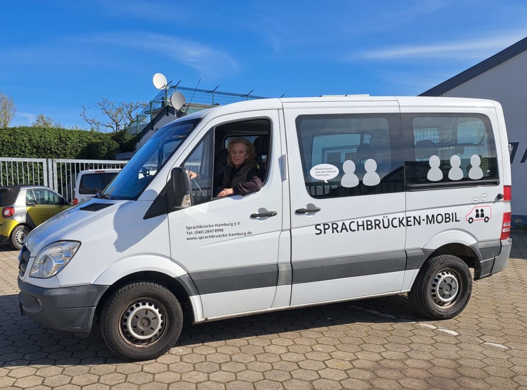 SB-Mobil_Annja als Fahrerin_2024-04-17 Ein weißer Kleintransporter seitlich steht auf einem großen Platz. Am Fahrersitz sitz eine Frau und lächelt in die Kamera