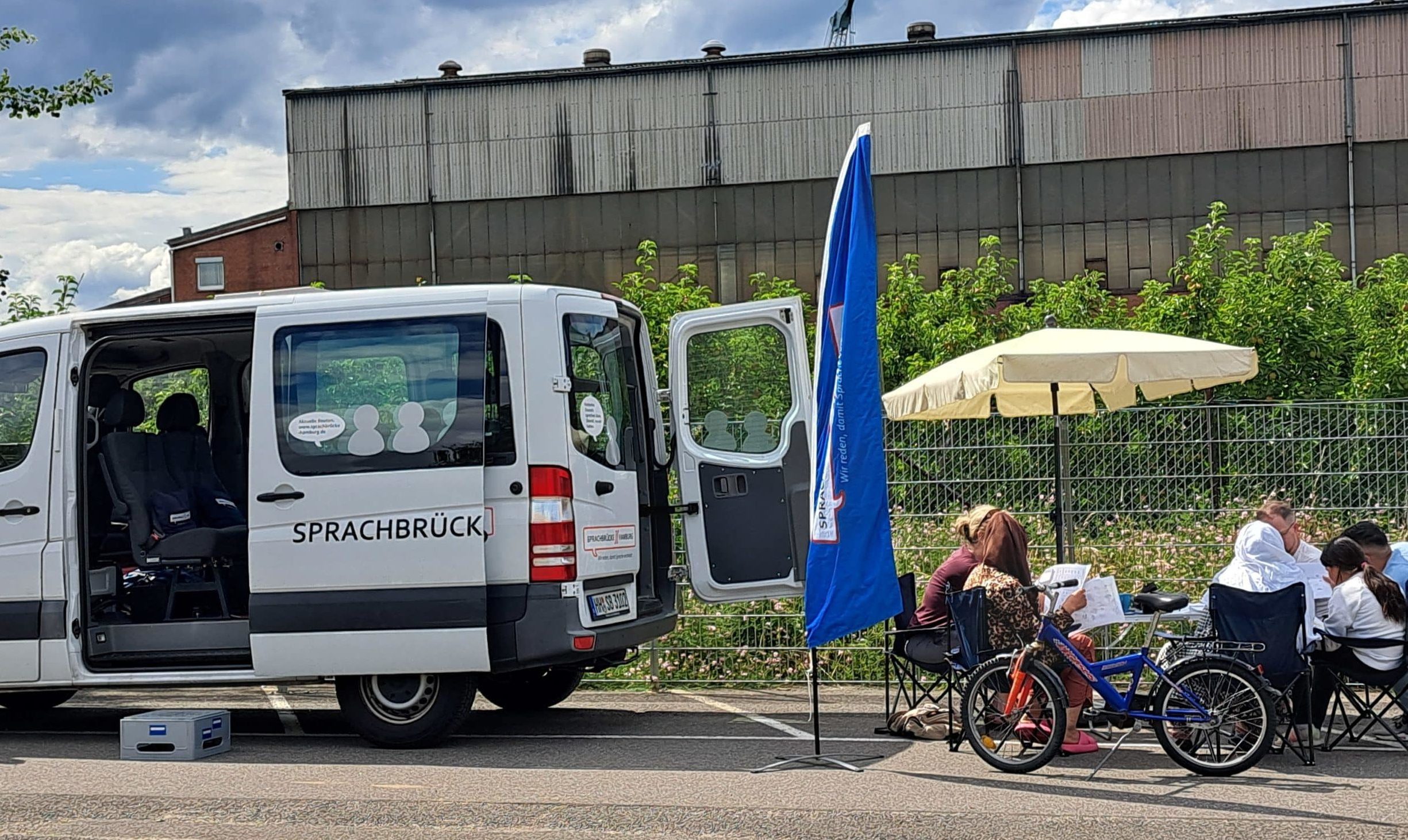 SB-Mobil_mobile GR_in Neuenfelde_2024-07-15 Ein weißer Kleintransporter steht mit offener Seiten- und HIntertür auf einem großen Platz. Daneben sitzen Menschen unter einem Sonnenschirm im Stuhlkreis und unterhalten sich.